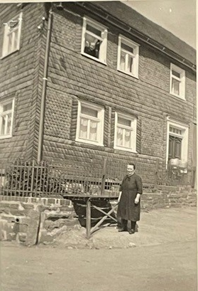 Schneiders Tante vor einem Milchbock bei Lotzes. Im Hintergrund bei der Haustür zum Trocknen aufgehängte Milchgefäße.